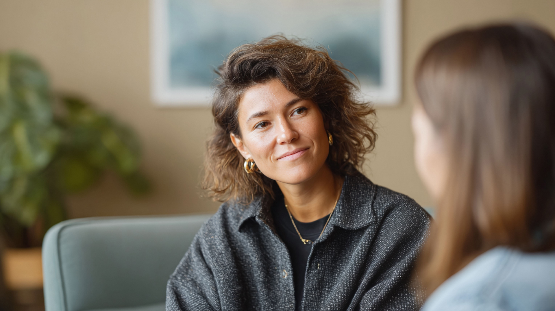 A woman with wavy brown hair and gold hoop earrings listens attentively to another person in an indoor setting.