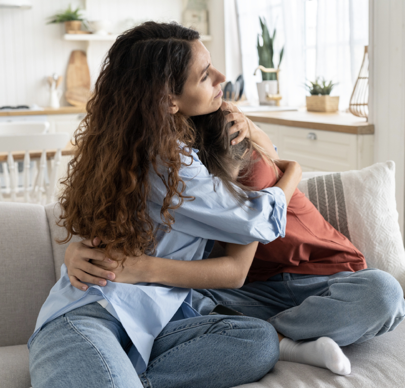 Who Do We Work With_ A woman and a child sit on a couch in a kitchen, hugging each other closely.