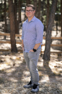 Man wearing glasses, a light blue button-up shirt, grey jeans, and sneakers stands outdoors in a wooded area with trees and a wooden fence in the background.