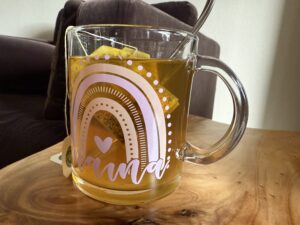 A clear glass mug with “mama” and a rainbow design holds yellow tea with a tea bag and metal spoon, placed on a wooden table near a couch.