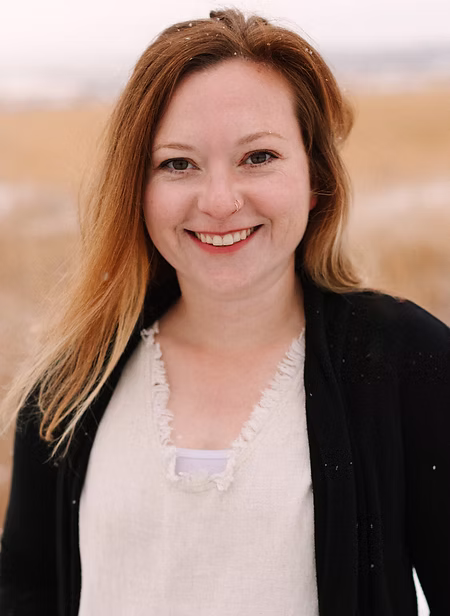 Nov2023-57.jpg A woman with light brown hair and a nose ring smiles at the camera, standing outdoors in a field with a light dusting of snow on her shoulders.