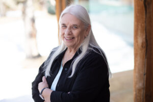Older woman with long white hair, wearing a black shirt, stands indoors near a wooden post, smiling at the camera with arms crossed.