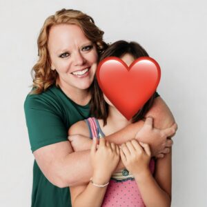 A smiling woman in a green shirt hugs a young girl whose face is covered with a large red heart emoji, against a plain white background.